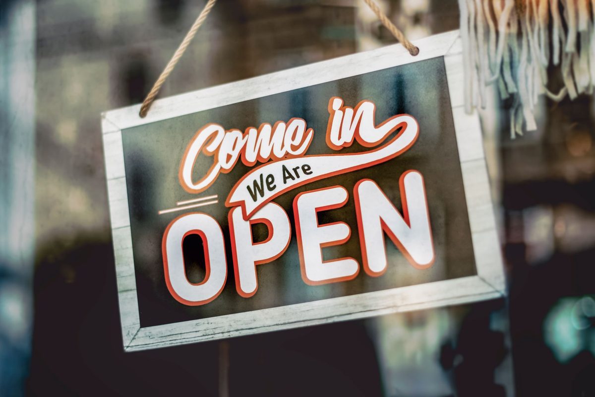 A framed sign reading Come in, We Are OPEN hangs by rope in a shop window