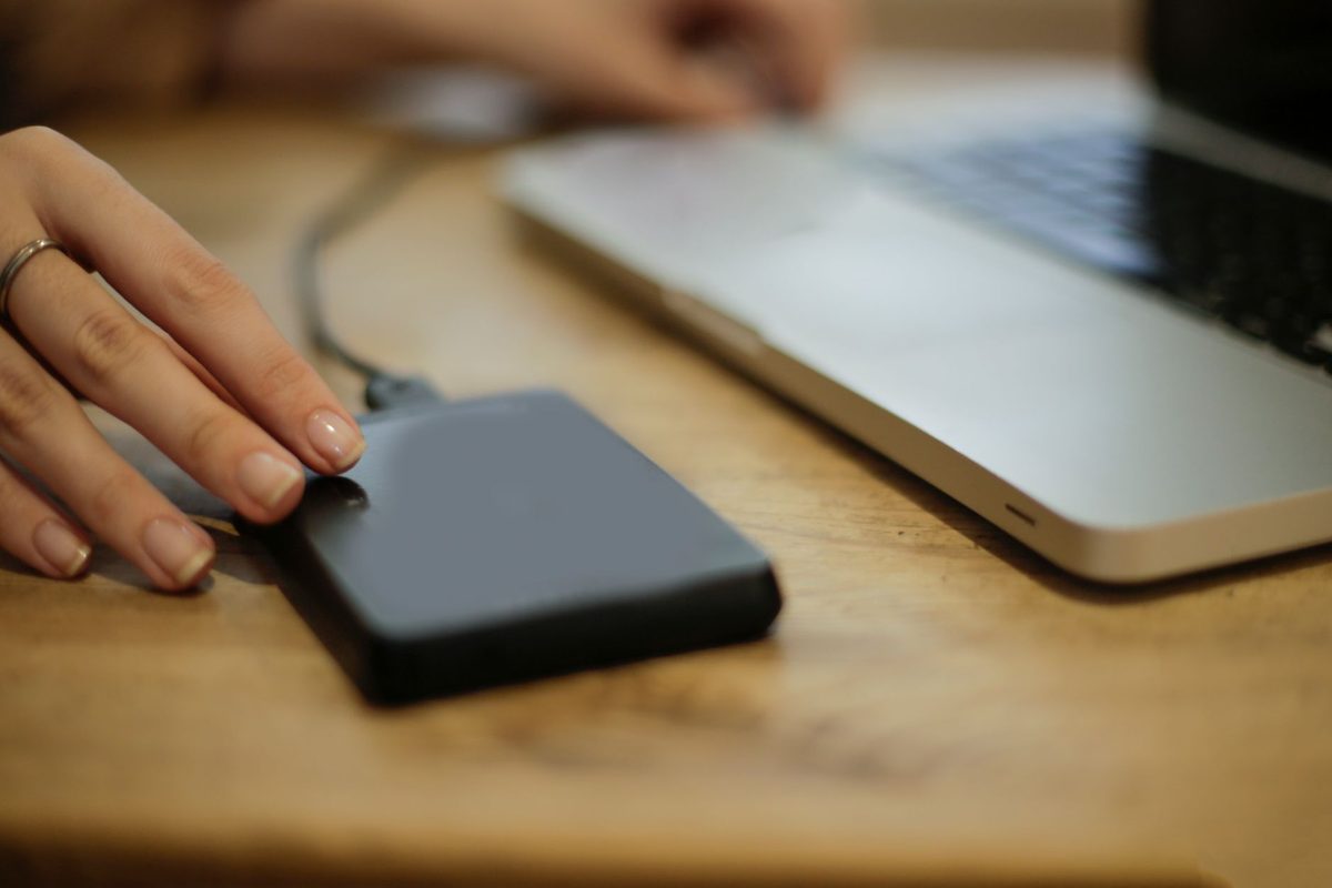 A hand rests on a portable external hard drive beside a silver laptop on a wooden desk